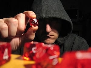 Image shows someone in a black hood holding a die, with a table covered in dice in the foreground. 