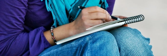 Image shows someone writing in a notebook that's rested on their knees.