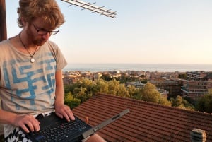 Image shows a young man sitting on a roof, working on his laptop. 
