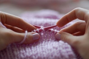Image shows someone knitting with red needles and pink wool.