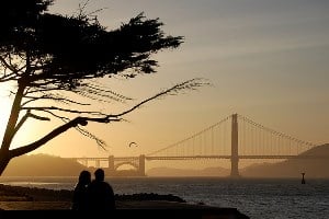 Image shows a couple sharing a romantic moment in front of a bridge by the sunset. 