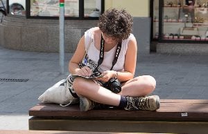 Image shows a young woman with a camera around her neck writing in a notebook. 